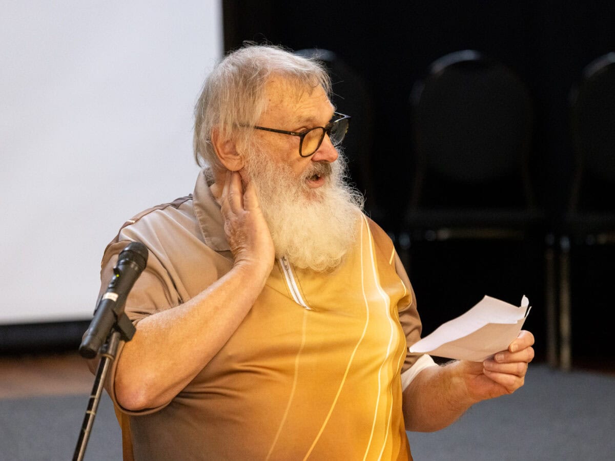 Elderly man with white beard and glasses speaking at a public event in Paekākāriki, New Zealand, holding papers and standing next to a microphone, representing local community engagement or storytelling in a scenic coastal town.